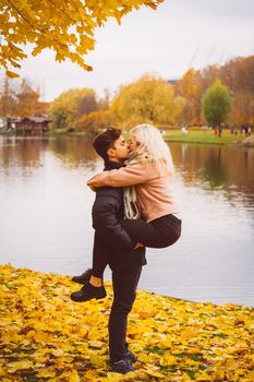 Cute brunette guy holds in his arms and kisses a beautiful blonde girl. Loving teens are happy, smiling, kissing. The concept of teenage love walking outdoors