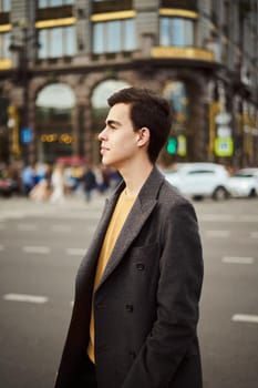 Handsome stylish fashionable man, brunette in an elegant gray coat, stands on street in historical center of St. Petersburg. Young man with dark hair, thick eyebrows.