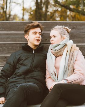 Teenagers in love are sitting on a park bench in the autumn, chatting cheerfully, chatting. Teenage love concept. Cute brunette and beautiful blonde are happy, smiling, laughing