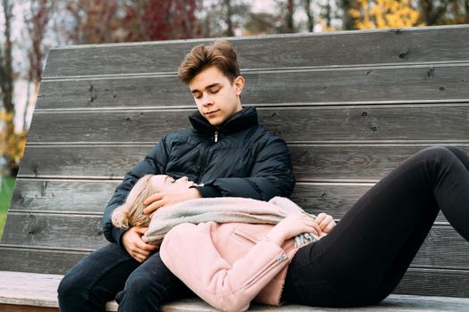 Teenagers in love are sitting on park bench in a autumn, chatting cheerfully, talking. Cute blonde girl lies, her head on boy lap. Teenage love concept.