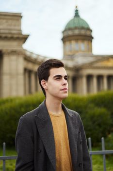 Handsome stylish fashionable man, a brunette in an elegant gray coat, stands on a street in the historical center of St. Petersburg. Young man with dark hair, thick eyebrows.