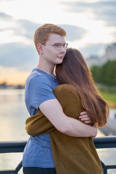 Girl with long thick dark hear embracing redhead boy in the blue t-shirt on bridge, teen love at sunset, summer time