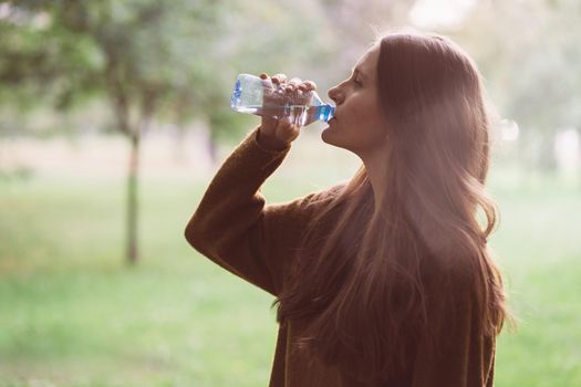 Young beautiful girl drinking water from a plastic bottle on the street in the Park in autumn or winter. A woman with beautiful long thick dark hair quenches her thirst for water on a walk, lens flare, light leak filter,