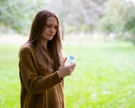 Young beautiful girl drinking water from a plastic bottle on the street in the Park in autumn or winter. A woman with beautiful long thick dark hair looks at a bottle of water and examines the composition of the liquid
