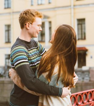 Girl with long thick dark hear embracing redhead boy in the blue t-shirt on bridge, young couple. Concept of teenage love and first kiss, sincere feelings of man and woman, city, waterfront. Vertical