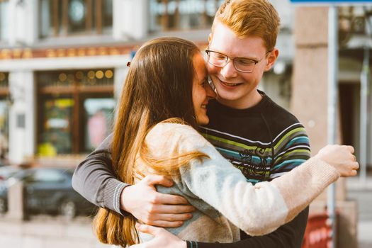 A red-haired young man embraces a young woman with long dark hair, they laugh and fool around, standing on a bridge in the center of the city. Concept of first teenage love, serious relationship, love and trust, happiness