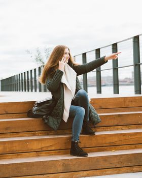 Beautiful young woman in warm clother is talking on phone and pointing with her hand. Girl sits on the wooden steps on waterfront in port in autumn, winter, vertical