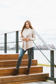 Beautiful young long-haired Caucasian woman stands on steps of wooden platform on waterfront at pier in port, vertical