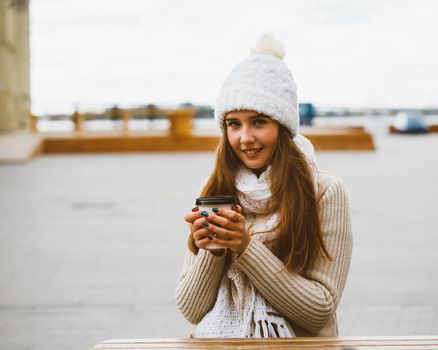 Beautiful young girl looking at camera, holding coffee, tea in reusable plastic mug in autumn, winter. A woman in warm clothes sitting on waterfront on Baltic sea in port, warmed by a hot drink