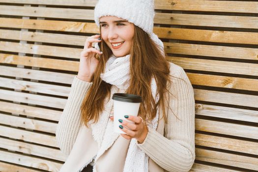 Beautiful young girl drinking coffee, tea from a plastic mug in autumn, winter and talking on a mobile phone. Woman with long hair sitting on a bench in autumn or winter, basking in a hot drink, copy space