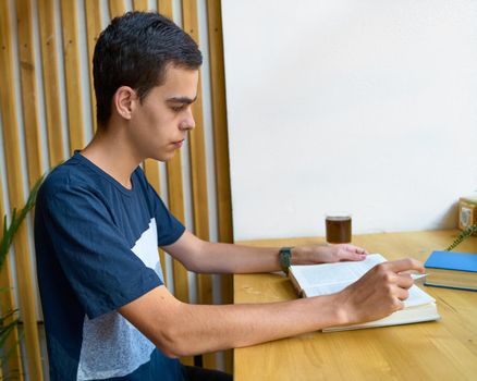 Young man with black hair reading a book, teenager in casual clothes in library, copy space