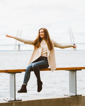 Happy young girl sitting on the waterfront at pier in port, enjoying life and waving his arms. Woman with long hair smiles and enjoys the moment