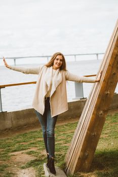 Happy young girl standing on the waterfront at pier in port, enjoying life and waving his arms. Woman with long hair smiles and enjoys moment, vertical