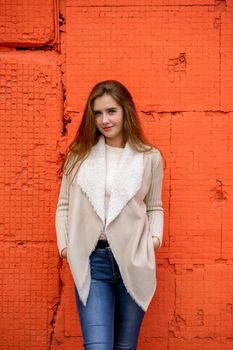 A beautiful girl stands against a bright wall of orange coral color, a woman with long hair in warm clothes on a colorful colored background