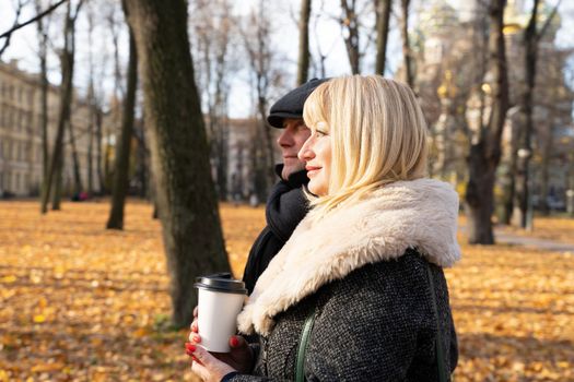 Happy blonde mature woman and handsome middle-aged brunette man walk in park and drink coffee from a reusable cup. A loving couple of 45-50 years old walks in autumn park in warm clothes