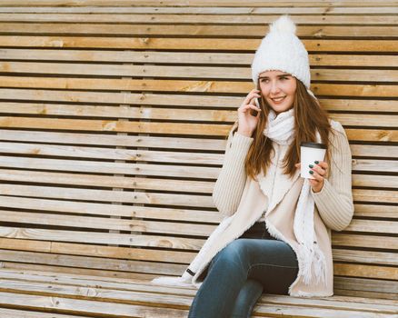 Beautiful young girl drinking coffee, tea from a plastic mug in autumn, winter and talking on a mobile phone. Woman with long hair sitting on a bench in autumn or winter, basking in a hot drink, copy space, close up