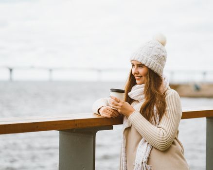 Beautiful young girl drinking coffee, tea from plastic mug in autumn, winter. A woman with long hair stands on waterfront on Baltic sea in port and waiting for ferry, heated by a hot drink, copy space