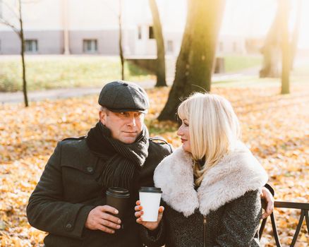 Happy blonde mature woman and handsome middle-aged brunette man sit on bench and drink coffee from a refillable cup. A loving couple of 45-50 years old walks in autumn or winter park in warm clothes