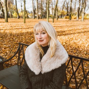 Happy blonde mature woman is sitting on bench in autumn park and thinking. Beautiful woman is relaxing in nature on sunny day. Portrait of a middle aged woman smiling and daydreaming.