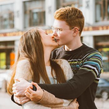 The redhead boy looks tenderly at girl and kiss. Concept of teenage love and first kiss, love, relationship, couple. City, waterfront.