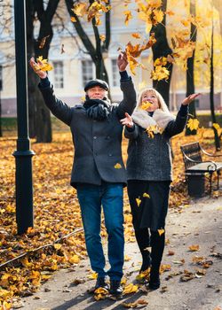 Happy blonde mature woman and handsome middle-aged brunette man walk in park and toss leaves. A loving couple of 45-50 years old walks in autumn park in warm clothes, vertical