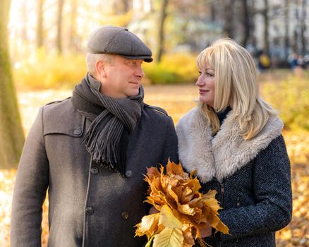 Happy blonde mature woman and handsome middle-aged brunette man walk in park, looking at each other. A loving couple of 45-50 years old walks in autumn park, holding bouquet of leaves
