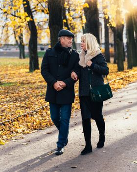 Happy blonde mature woman and handsome middle-aged brunette man walk in park, looking at each other. A loving couple of 45-50 years old walks in autumn park in warm clothes, vertical