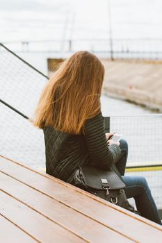 Unrecognizable woman with long hair sits with phone in her hand, her back to camera. Autumn or winter, a girl in jacket outdoors waiting for a call or sms, texting, vertical