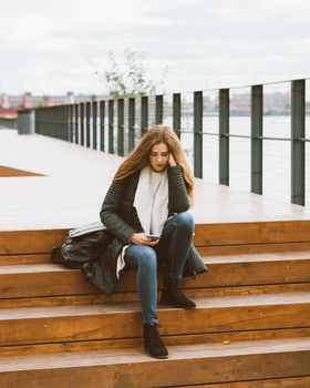Beautiful woman with long hair sits on stairs with phone in her hand, back to camera. Autumn or winter, girl in jacket outdoors waiting for call or sms, texting, vertical