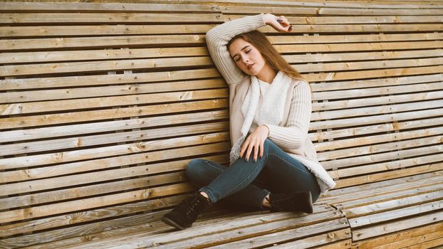 Beautiful young girl with long brown hair sits on a wooden bench made of planks and rests, relaxes and reflects. Outdoor photo shoot with attractive woman in winter or autumn, banner