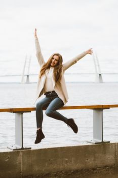 Happy young girl sitting on the waterfront at pier in port, enjoying life and waving his arms. Woman with long hair smiles and enjoys the moment