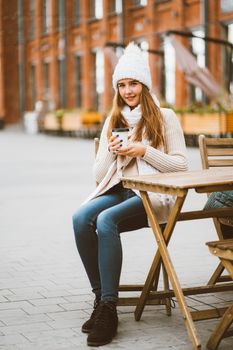 Beautiful young girl drinking coffee, tea from plastic reusable mug in autumn, winter. Woman with long hair, in warm clothes sitting in a street cafe, warmed by hot drink, vertical