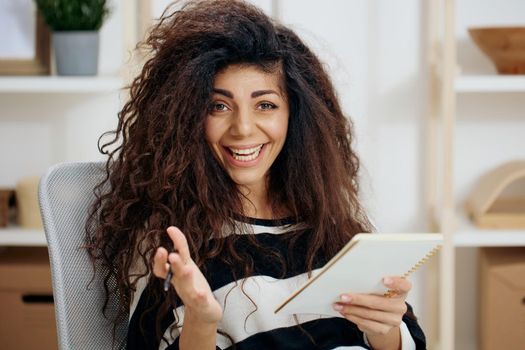 Joyful smiling happy pretty beautiful curly tanned Latin woman in striped shirt hold notebook look at camera say Yeah write work letter essay in home interior. Copy space Banner. Writer concept
