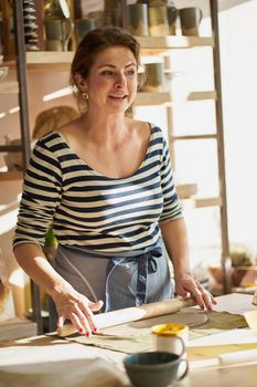 Beautiful happy woman making ceramic ware in workplace, smiling in sun light. Concept for woman in freelance, business, hobby