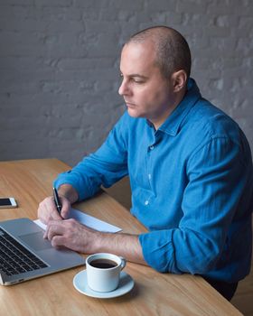 Handsome handsome mature man writes on a sheet of paper and looks at the computer screen, laptop. Man with casual clothes in a blue shirt at a table in the office in front of the window, vertical