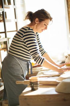 Beautiful happy woman making ceramic ware in workplace, laughing, smiling in sun light. Concept for woman in freelance, business, hobby