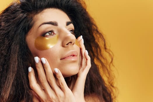 Pensive attractive Latin lady with gold hydrogel patches under eye touch soft skin look aside posing isolated over yellow background. Cosmetic product ad Natural beauty Skincare concept Copy space