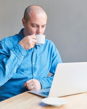 A handsome middle-aged man drinks coffee from a mug and looks at the screen of a computer, laptop. Man with casual wear in a blue shirt is sitting in the office. Vertical