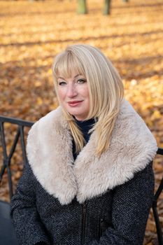 Happy blonde mature woman is sitting on a bench in the autumn park and thinking. Beautiful woman is relaxing in nature on sunny day. Portrait of middle aged woman smiling and daydreaming.