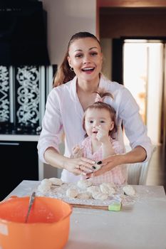 Portrait of gorgeous young adult mother in shirt cooking with her little daughter in similar pink plaid shirt. They are smiling while kneading dough in the kitchen.