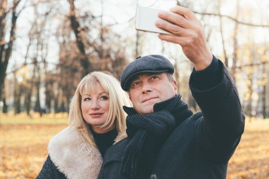 Happy blonde mature woman and beautiful middle-aged brunette take selfie on mobile phone. Loving couple of 45-50 years old walks in the autumn park in warm clothes, in coat and enjoys life