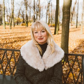 Happy blonde mature woman is sitting on a bench in the autumn park and thinking. Beautiful woman is relaxing in nature on sunny day. Portrait of middle aged woman smiling and daydreaming.