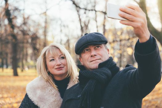 Happy blonde mature woman and beautiful middle-aged brunette take a selfie on the phone. Loving couple of 45-50 years old walks in the autumn park in warm clothes, in a coat and enjoys life