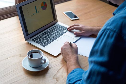 Unrecognizable man writes plan on paper and looks at charts on computer screen, laptop. A man with casual clothes in blue shirt sits in office in front of window, view from the back