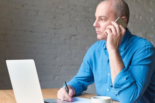 Handsome handsome mature man looks out window and talking on cell phone, sitting at computer, laptop. Man with casual clothes in a blue shirt and table at office in front of window, copy space