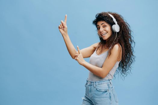 MUSIC STREAMING PLATFORM AD. Overjoyed pretty curly Latin female in headphones, listen funny song, smile, look at camera point fingers up, say Look at this. Cool offer over blue background.
