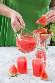 a woman in a green dress pours a red refreshing drink from the pulp of a watermelon into a glass glass, bright natural background