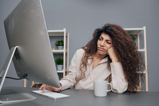 Unhappy tired tanned adorable curly Latin businesswoman in linen shirt recline on hand hate work time in home office. Copy space. Attractive freelancer work from home using modern desktop computer