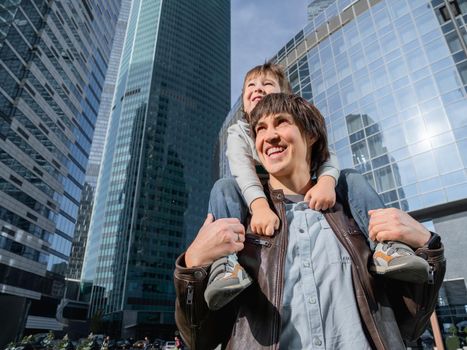 Little boy sits on father's shoulders among skyscrapers. Dad and son looks on glass walls of buildings. Future and modern technologies, life balance and family life in well keeps districts.