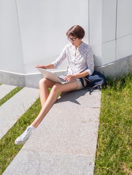 Woman sits with laptop on urban park bench. Freelancer at work. Student learns remotely from outdoors. Modern lifestyle. Summer vibes. Outdoor workplace.